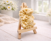 Load image into Gallery viewer, A detailed handmade vegan soap, sculpted as a cheerful pig sitting in a mound of bubbles and holding a scrubbing brush, displayed on a miniature wooden easel against a soft farmhouse window background 
