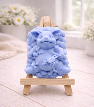 Load image into Gallery viewer, A detailed handmade vegan soap, sculpted as a cheerful pig sitting in a mound of bubbles and holding a scrubbing brush, displayed on a miniature wooden easel against a soft farmhouse window background 

