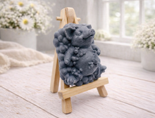 Load image into Gallery viewer, A detailed handmade vegan soap, sculpted as a cheerful pig sitting in a mound of bubbles and holding a scrubbing brush, displayed on a miniature wooden easel against a soft farmhouse window background 
