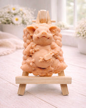 Load image into Gallery viewer, A detailed handmade vegan soap in a natural cream tone, sculpted as a cheerful pig sitting in a mound of bubbles and holding a scrubbing brush, displayed on a miniature wooden easel against a soft farmhouse window background.

