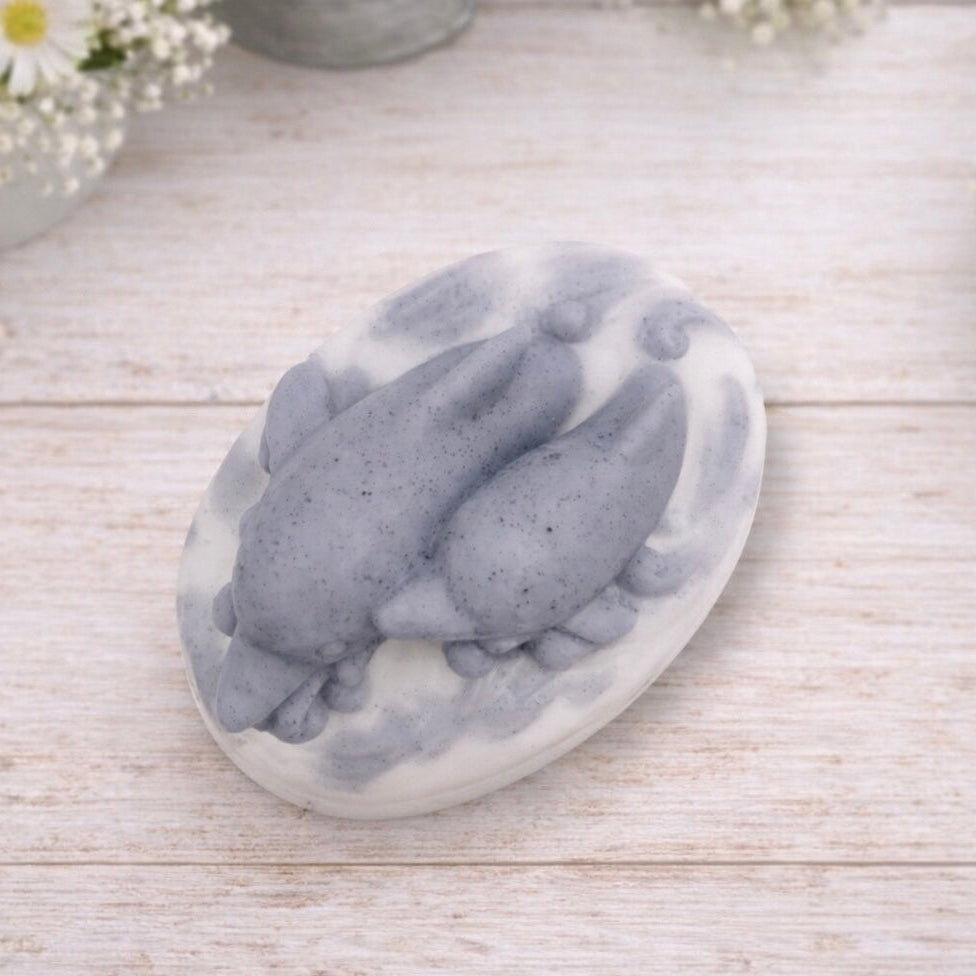 A handmade vegan soap featuring two grey dolphins jumping through white frothy waves; the base is white, and the dolphins are highly detailed, displayed on a light wooden surface against a soft farmhouse background.