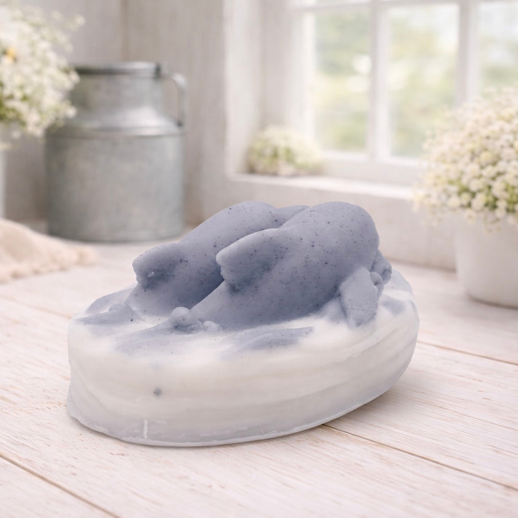 A handmade vegan soap featuring two grey dolphins jumping through white frothy waves; the base is white, and the dolphins are highly detailed, displayed on a light wooden surface against a soft farmhouse background.