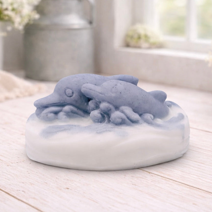 A handmade vegan soap featuring two grey dolphins jumping through white frothy waves; the base is white, and the dolphins are highly detailed, displayed on a light wooden surface against a soft farmhouse background.
