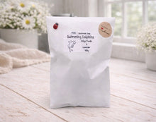 Load image into Gallery viewer, White bag labeled 'Swimming Dolphins' on a wooden surface with flowers in the background
