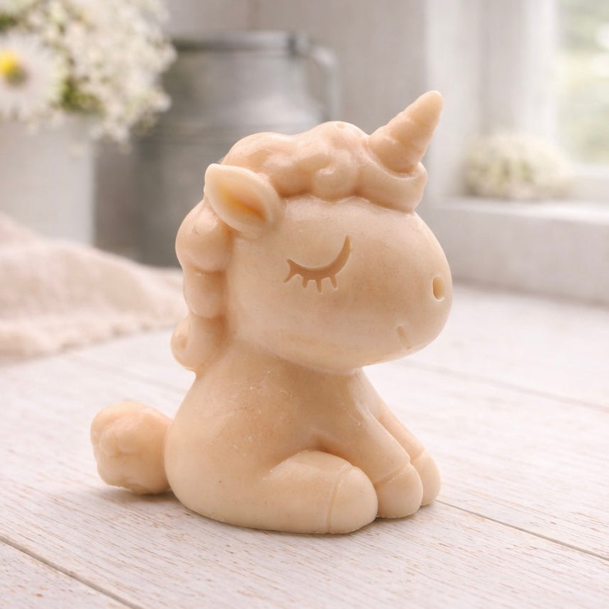 A 3D handmade vegan soap sculpted into a cute sitting unicorn with a spiralled horn, a smiling expression with a closed winking eye, and a bubbly mane and tail, displayed on a light wooden surface in a bright farmhouse setting.