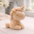 A 3D handmade vegan soap sculpted into a cute sitting unicorn with a spiralled horn, a smiling expression with a closed winking eye, and a bubbly mane and tail, displayed on a light wooden surface in a bright farmhouse setting.