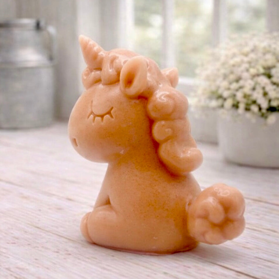 A 3D handmade vegan soap sculpted into a cute sitting unicorn with a spiralled horn, a smiling expression with a closed winking eye, and a bubbly mane and tail, displayed on a light wooden surface in a bright farmhouse setting.