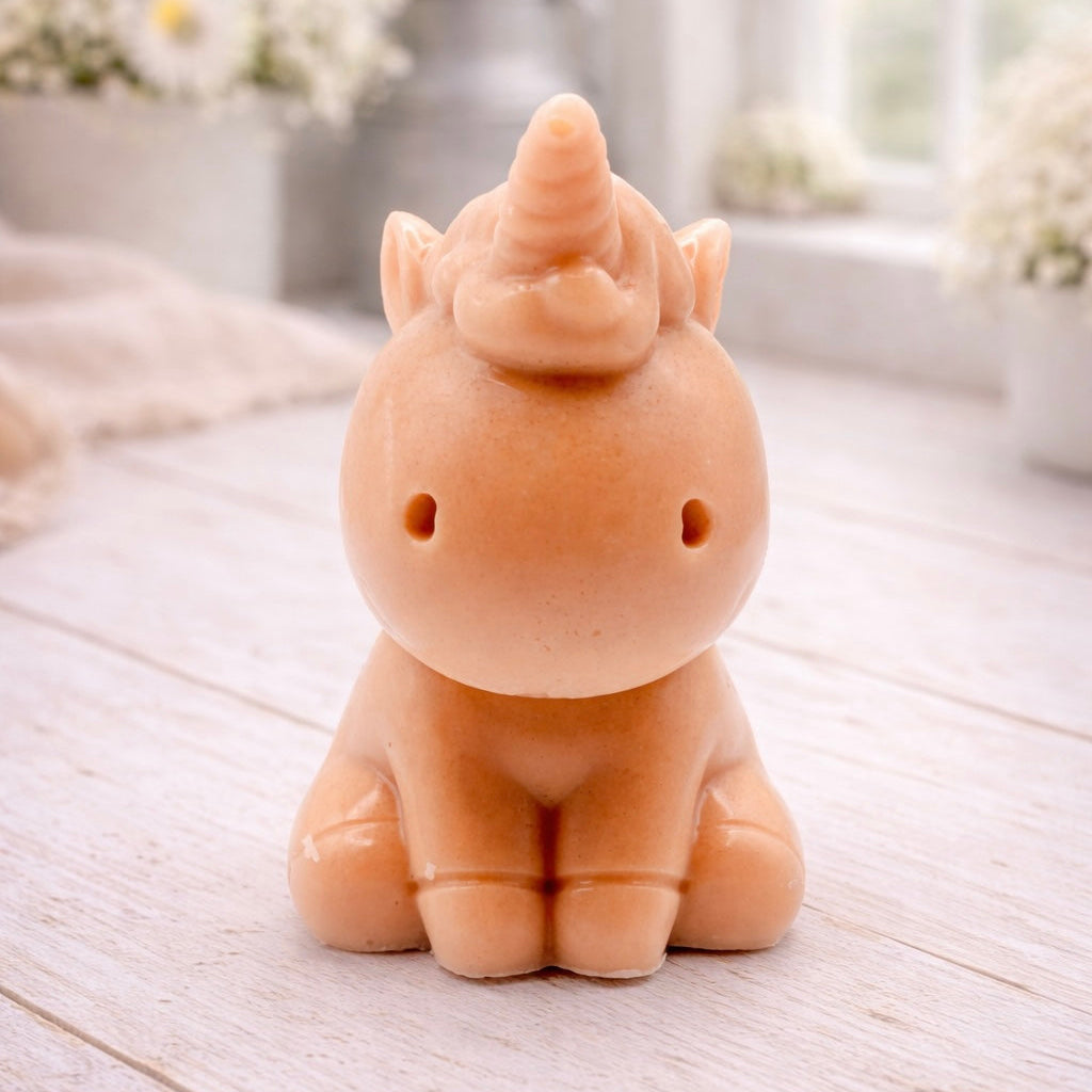 A 3D handmade vegan soap sculpted into a cute sitting unicorn with a spiralled horn, a smiling expression with a closed winking eye, and a bubbly mane and tail, displayed on a light wooden surface in a bright farmhouse setting.