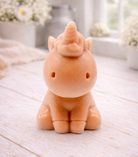 Load image into Gallery viewer, A 3D handmade vegan soap sculpted into a cute sitting unicorn with a spiralled horn, a smiling expression with a closed winking eye, and a bubbly mane and tail, displayed on a light wooden surface in a bright farmhouse setting.
