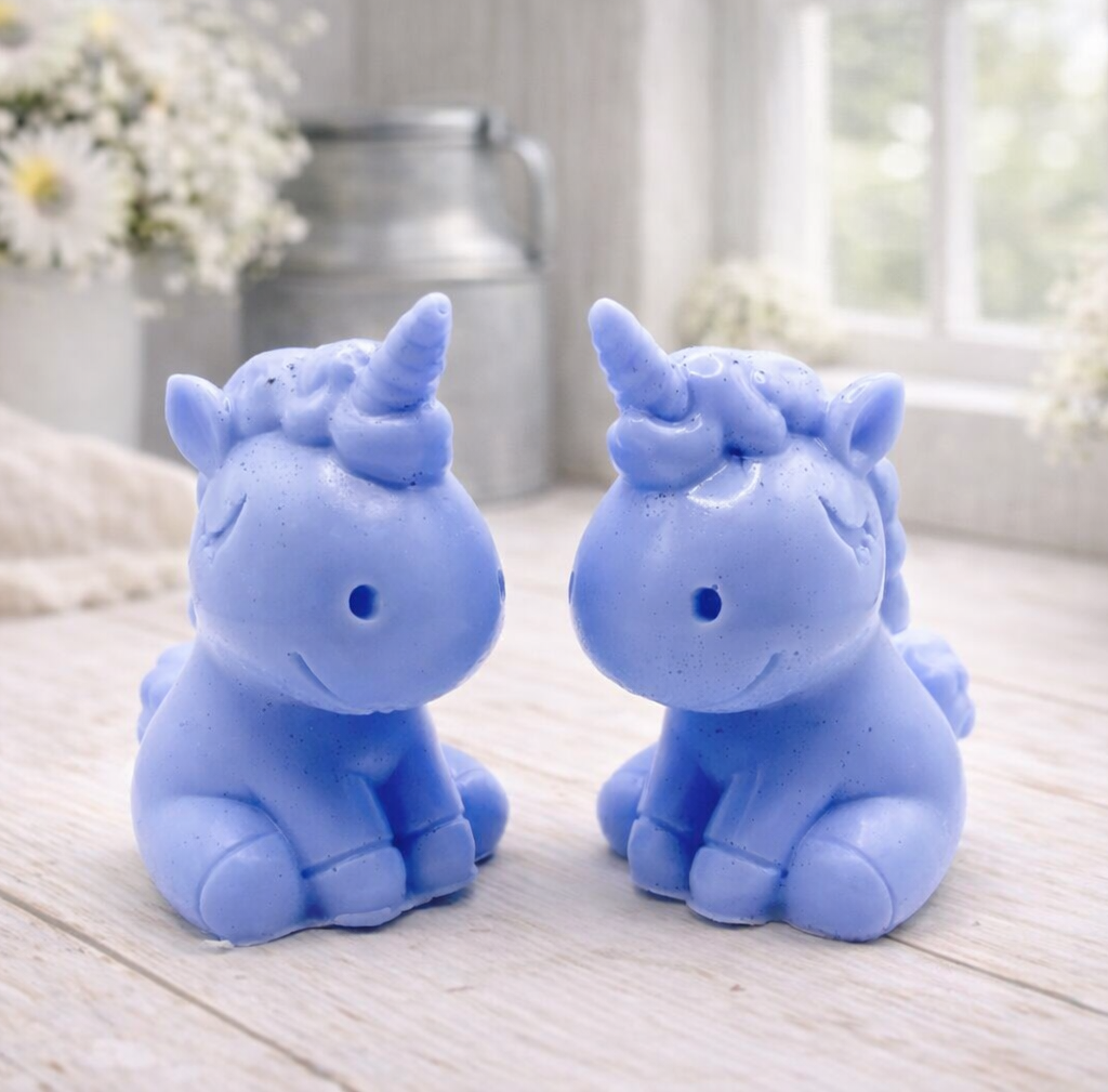 A 3D handmade vegan soap sculpted into a cute sitting unicorn with a spiralled horn, a smiling expression with a closed winking eye, and a bubbly mane and tail, displayed on a light wooden surface in a bright farmhouse setting.
