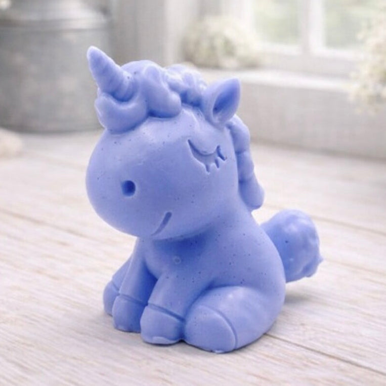 A 3D handmade vegan soap sculpted into a cute sitting unicorn with a spiralled horn, a smiling expression with a closed winking eye, and a bubbly mane and tail, displayed on a light wooden surface in a bright farmhouse setting.