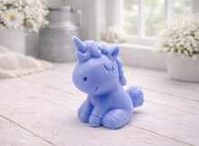 Load image into Gallery viewer, A 3D handmade vegan soap sculpted into a cute sitting unicorn with a spiralled horn, a smiling expression with a closed winking eye, and a bubbly mane and tail, displayed on a light wooden surface in a bright farmhouse setting.
