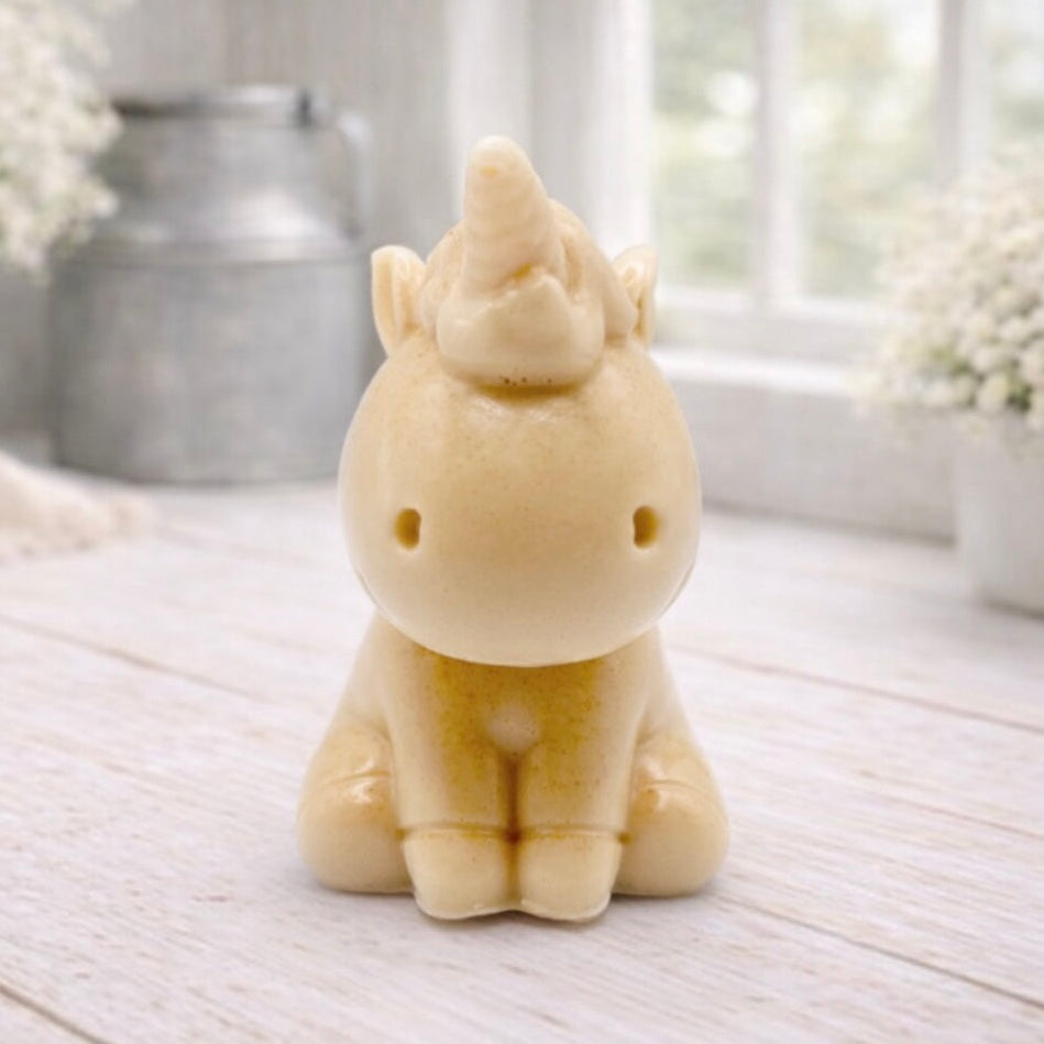 A 3D handmade vegan soap sculpted into a cute sitting unicorn with a spiralled horn, a smiling expression with a closed winking eye, and a bubbly mane and tail, displayed on a light wooden surface in a bright farmhouse setting.