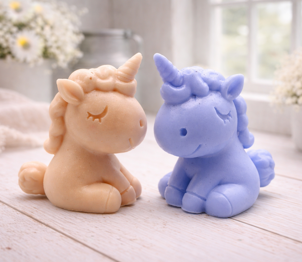 A handmade vegan soap sculpted into a cute sitting unicorn with a spiralled horn, a smiling expression with a closed winking eye, and a bubbly mane and tail, displayed on a light wooden surface in a bright farmhouse setting.