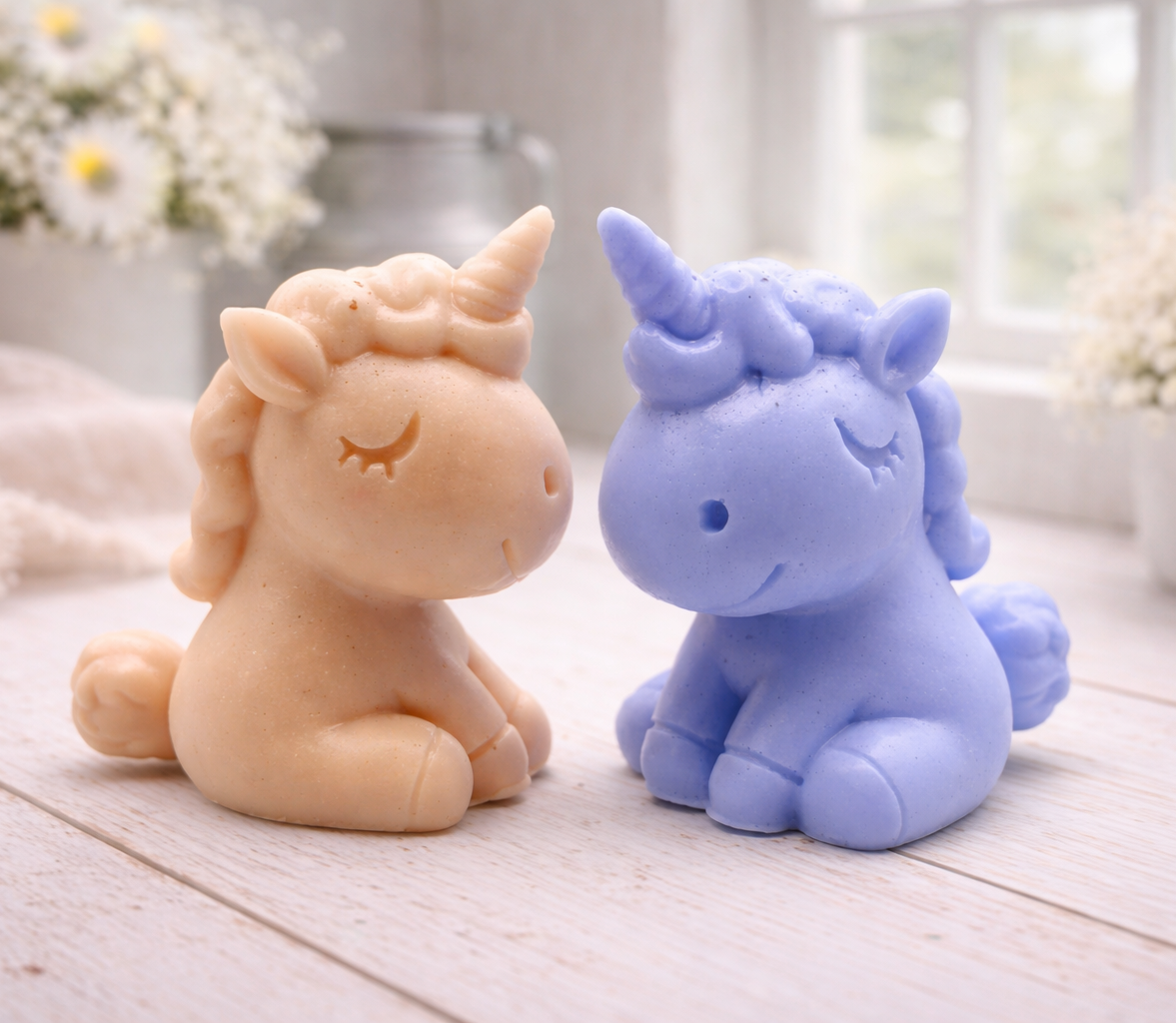 A handmade vegan soap sculpted into a cute sitting unicorn with a spiralled horn, a smiling expression with a closed winking eye, and a bubbly mane and tail, displayed on a light wooden surface in a bright farmhouse setting.