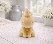 Load image into Gallery viewer, A 3D handmade vegan soap sculpted into a cute sitting unicorn with a spiralled horn, a smiling expression with a closed winking eye, and a bubbly mane and tail, displayed on a light wooden surface in a bright farmhouse setting.
