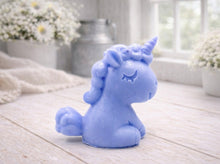 Load image into Gallery viewer, A 3D handmade vegan soap sculpted into a cute sitting unicorn with a spiralled horn, a smiling expression with a closed winking eye, and a bubbly mane and tail, displayed on a light wooden surface in a bright farmhouse setting.
