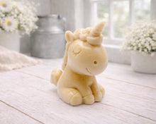 Load image into Gallery viewer, A 3D handmade vegan soap sculpted into a cute sitting unicorn with a spiralled horn, a smiling expression with a closed winking eye, and a bubbly mane and tail, displayed on a light wooden surface in a bright farmhouse setting.
