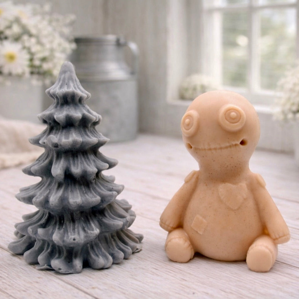 A handmade vegan soap gift set featuring a cream-coloured voodoo doll soap with stitched details and a grey tiered Christmas tree soap, presented in a black gift box tied with a red raffia ribbon and a small ladybird embellishment.