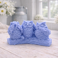 A handmade vegan soap gift featuring three owls perched on a branch in the "See No Evil, Hear No Evil, Speak No Evil" poses, set against a bright window with white flowers.