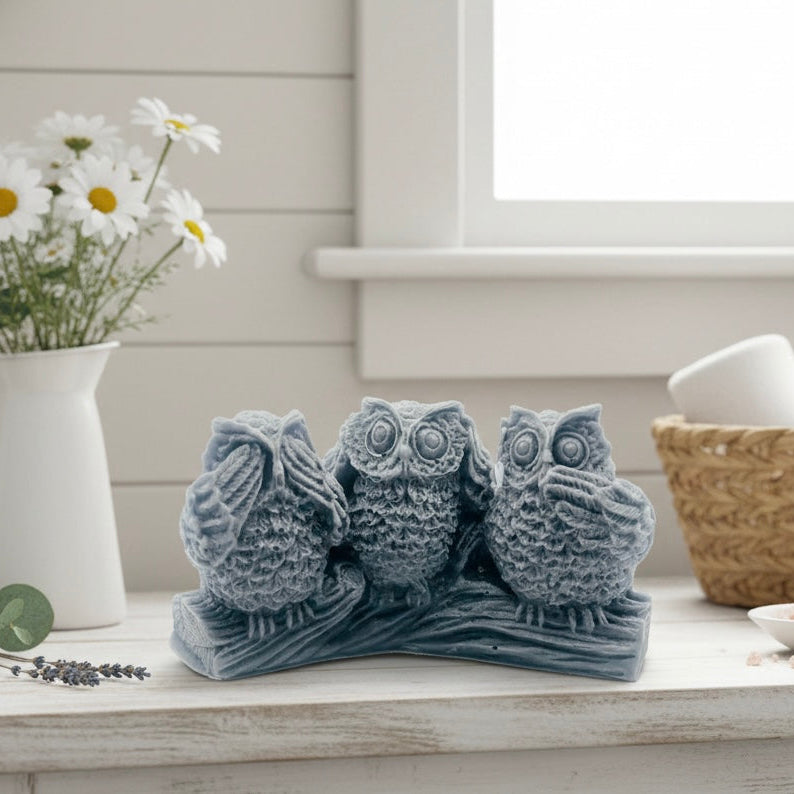 A set of three detailed handmade vegan owl soaps in a "See No, Hear No, Speak No Evil" pose, displayed on a rustic log base with a soft pastel farmhouse decor background.