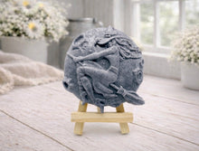 Load image into Gallery viewer, A circular handmade vegan soap featuring a detailed high-relief carving of a witch wearing a pointed hat, flying on a broomstick across a cratered moon, displayed on a miniature wooden easel.
