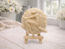 Load image into Gallery viewer, A circular handmade vegan soap featuring a detailed high-relief carving of a witch wearing a pointed hat, flying on a broomstick across a cratered moon, displayed on a miniature wooden easel.
