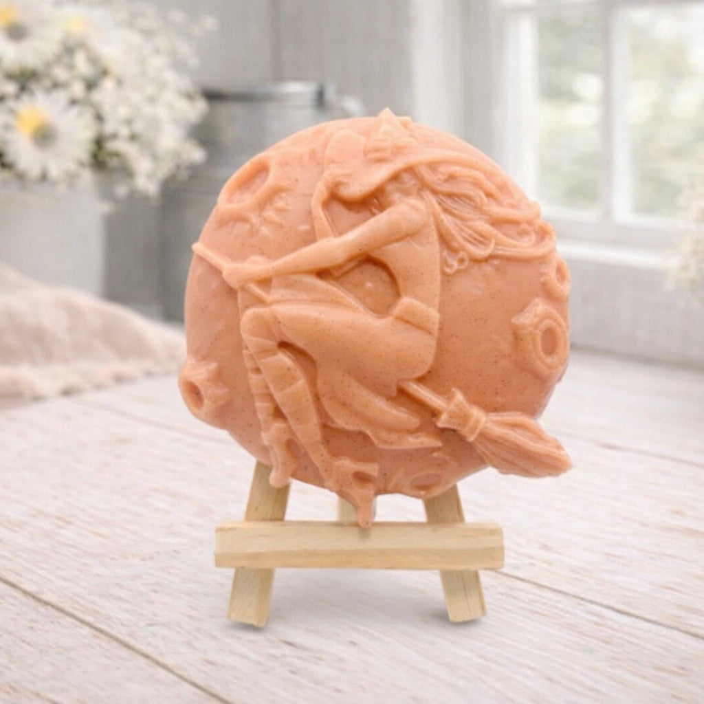 A circular handmade vegan soap featuring a detailed high-relief carving of a witch wearing a pointed hat, flying on a broomstick across a cratered moon, displayed on a miniature wooden easel.