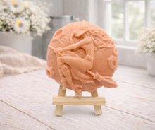 Load image into Gallery viewer, A circular handmade vegan soap featuring a detailed high-relief carving of a witch wearing a pointed hat, flying on a broomstick across a cratered moon, displayed on a miniature wooden easel.
