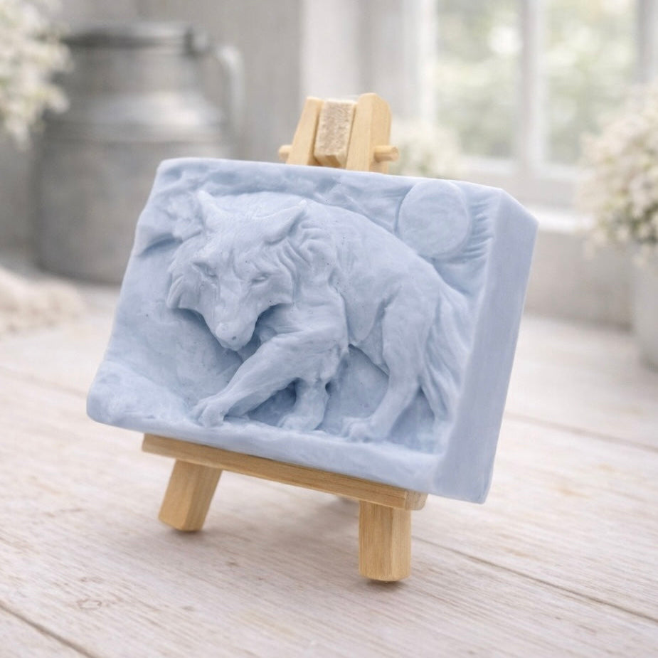 A rectangular handmade vegan soap, featuring a detailed relief carving of a wolf in mid-stride across a rugged landscape with a large full moon in the background, displayed on a miniature wooden easel.