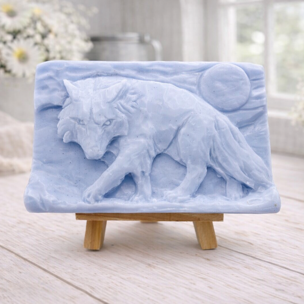 A rectangular handmade vegan soap, featuring a detailed relief carving of a wolf in mid-stride across a rugged landscape with a large full moon in the background, displayed on a miniature wooden easel.