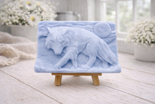 Load image into Gallery viewer, A rectangular handmade vegan soap, featuring a detailed relief carving of a wolf in mid-stride across a rugged landscape with a large full moon in the background, displayed on a miniature wooden easel.
