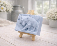 Load image into Gallery viewer, A rectangular handmade vegan soap, featuring a detailed relief carving of a wolf in mid-stride across a rugged landscape with a large full moon in the background, displayed on a miniature wooden easel.
