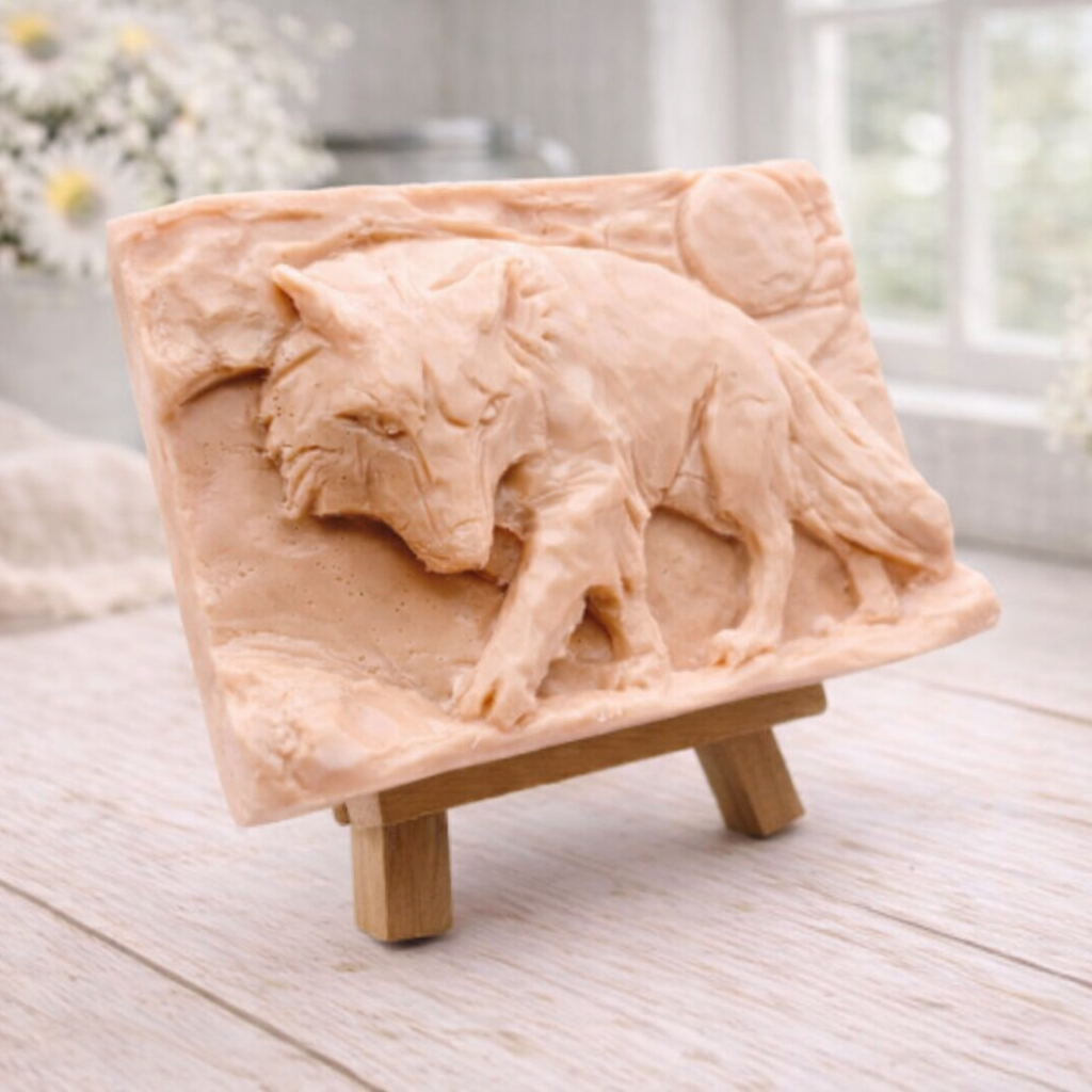 A rectangular handmade vegan soap, featuring a detailed relief carving of a wolf in mid-stride across a rugged landscape with a large full moon in the background, displayed on a miniature wooden easel.