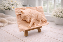 Load image into Gallery viewer, A rectangular handmade vegan soap, featuring a detailed relief carving of a wolf in mid-stride across a rugged landscape with a large full moon in the background, displayed on a miniature wooden easel.
