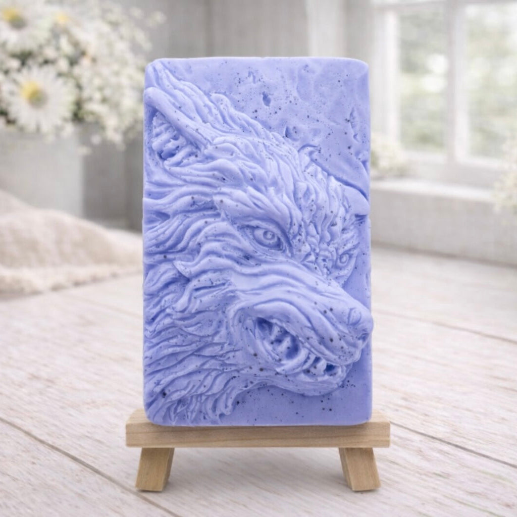 A rectangular handmade vegan soap featuring a highly detailed, 3D relief carving of a wolf's head in profile with a thick, textured mane, displayed on a miniature wooden easel.