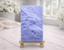 Load image into Gallery viewer, A rectangular handmade vegan soap featuring a highly detailed, 3D relief carving of a wolf's head in profile with a thick, textured mane, displayed on a miniature wooden easel.
