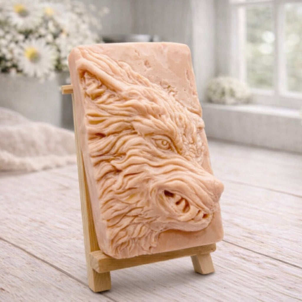 A rectangular handmade vegan soap featuring a highly detailed, 3D relief carving of a wolf's head in profile with a thick, textured mane, displayed on a miniature wooden easel.