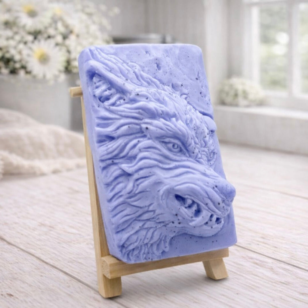 A rectangular handmade vegan soap featuring a highly detailed, 3D relief carving of a wolf's head in profile with a thick, textured mane, displayed on a miniature wooden easel.