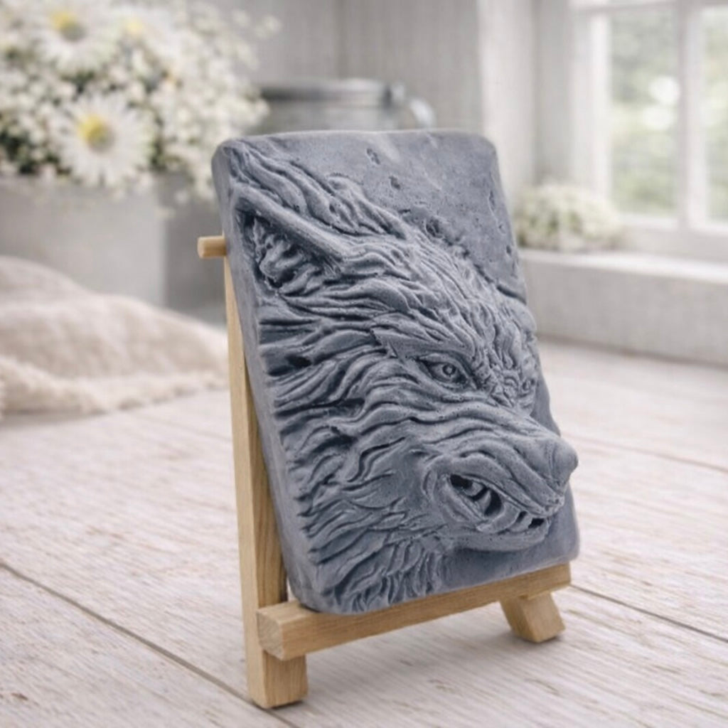 A rectangular handmade vegan soap featuring a highly detailed, 3D relief carving of a wolf's head in profile with a thick, textured mane, displayed on a miniature wooden easel.