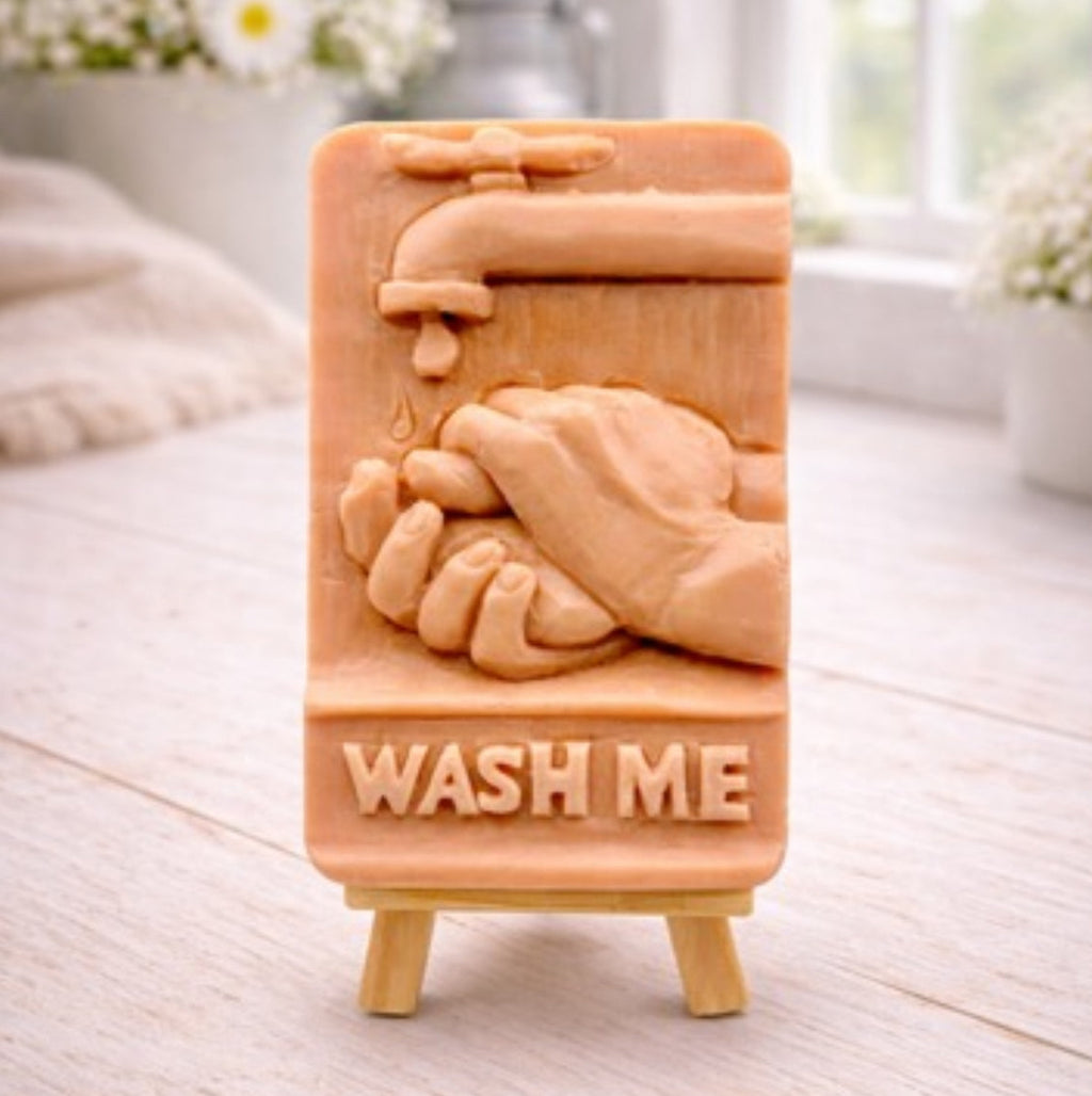 handmade vegan soap featuring a relief sculpture of hands being washed under a dripping tap with the words "WASH ME" at the base, displayed on a small wooden easel in a bright farmhouse decor kitchen setting.