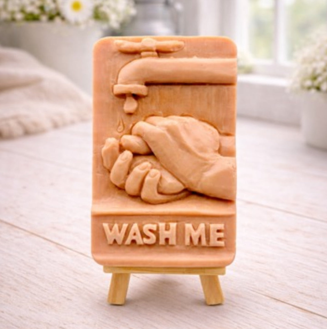 handmade vegan soap featuring a relief sculpture of hands being washed under a dripping tap with the words "WASH ME" at the base, displayed on a small wooden easel in a bright farmhouse decor kitchen setting.