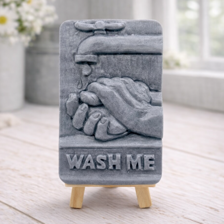 handmade vegan soap featuring a relief sculpture of hands being washed under a dripping tap with the words "WASH ME" at the base, displayed on a small wooden easel in a bright farmhouse decor kitchen setting.
