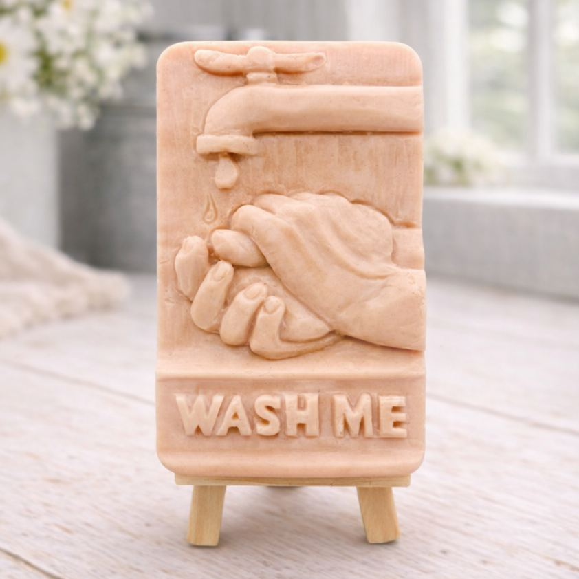 handmade vegan soap featuring a relief sculpture of hands being washed under a dripping tap with the words "WASH ME" at the base, displayed on a small wooden easel in a bright farmhouse decor kitchen setting.