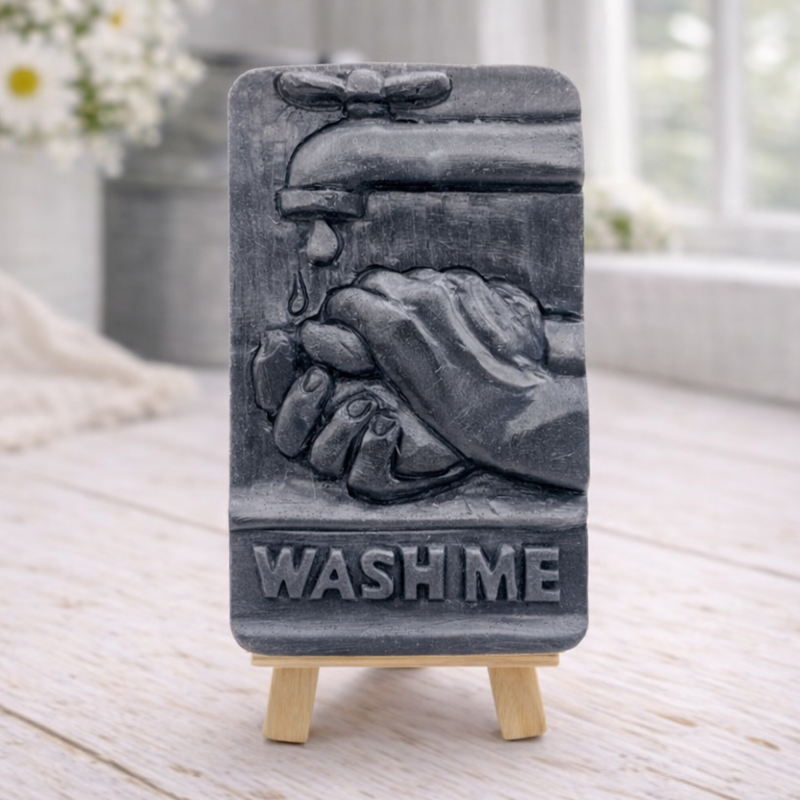 handmade vegan soap featuring a relief sculpture of hands being washed under a dripping tap with the words "WASH ME" at the base, displayed on a small wooden easel in a bright farmhouse decor kitchen setting.