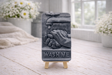 Load image into Gallery viewer, handmade vegan soap featuring a relief sculpture of hands being washed under a dripping tap with the words "WASH ME" at the base, displayed on a small wooden easel in a bright farmhouse decor kitchen setting.
