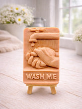 Load image into Gallery viewer, handmade vegan soap featuring a relief sculpture of hands being washed under a dripping tap with the words "WASH ME" at the base, displayed on a small wooden easel in a bright farmhouse decor kitchen setting.
