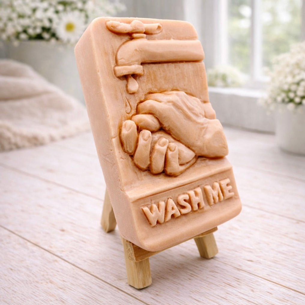 handmade vegan soap featuring a relief sculpture of hands being washed under a dripping tap with the words "WASH ME" at the base, displayed on a small wooden easel in a bright farmhouse decor kitchen setting.