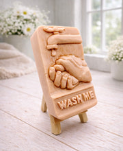 Load image into Gallery viewer, handmade vegan soap featuring a relief sculpture of hands being washed under a dripping tap with the words "WASH ME" at the base, displayed on a small wooden easel in a bright farmhouse decor kitchen setting.
